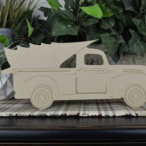 May include: A wooden cutout of a vintage pickup truck with a Christmas tree in the bed. The truck is painted a light brown color and is in profile view. The truck is on a plaid surface.