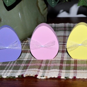 May include: Three pastel-colored Easter egg-shaped decorations. The eggs are lavender, pink, and yellow, each adorned with a sheer, white ribbon tied in a bow. They are arranged on a plaid tablecloth, suggesting a festive or seasonal theme.
