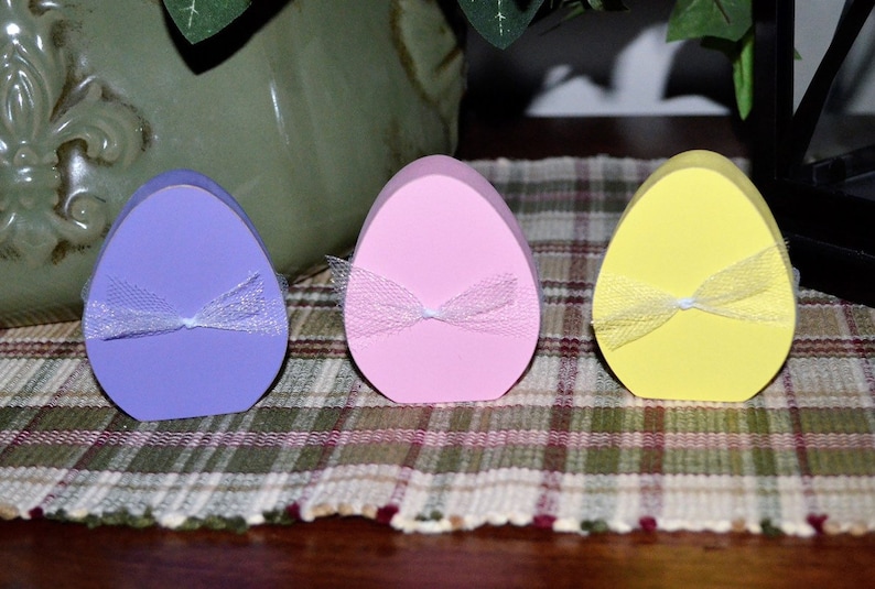 May include: Three pastel-colored Easter egg-shaped decorations. The eggs are lavender, pink, and yellow, each adorned with a sheer, white ribbon tied in a bow. They are arranged on a plaid tablecloth, suggesting a festive or seasonal theme.