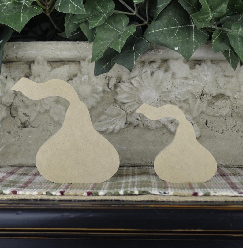 Op de afbeelding: Twee onafgewerkte houten peervormige vormen, een groter dan de andere, liggen op een geruit stofoppervlak. De peren zijn van hout en hebben een gladde, natuurlijke afwerking.
