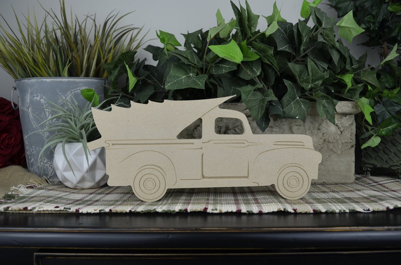 May include: A wooden cutout of a vintage pickup truck with a Christmas tree in the bed. The truck is painted in a natural wood color and is ready to be decorated.
