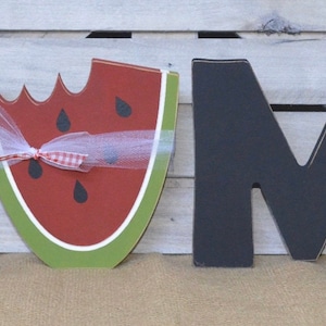 May include: Black wooden letters spelling "HOME" with a watermelon cutout in place of the "O". The watermelon is red with black seeds and has a green rind. A white ribbon with red polka dots is tied around the watermelon.
