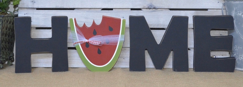 May include: Black wooden letters spelling "HOME" with a watermelon cutout in place of the "O". The watermelon is red with black seeds and has a green rind. A white ribbon with red polka dots is tied around the watermelon.
