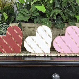 Op de afbeelding: Drie houten hartvormige decoraties met gouden strepen. De harten zijn rood, wit en roze.