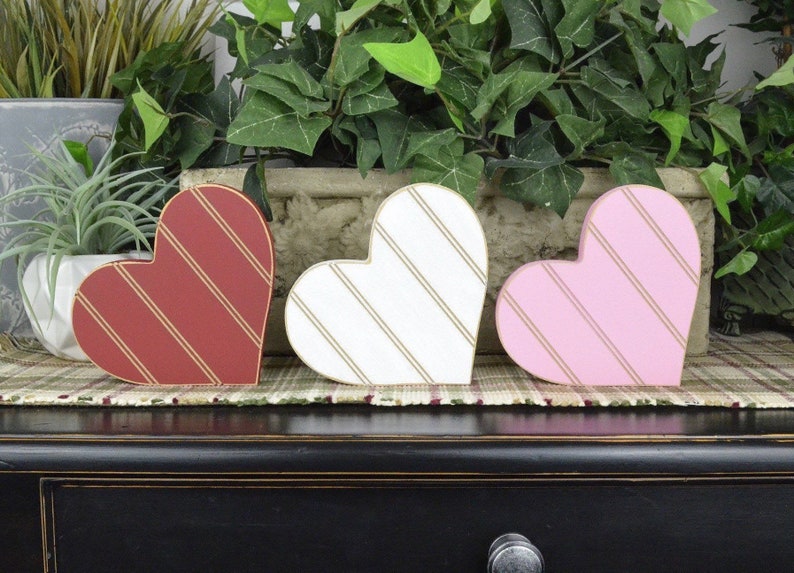 Op de afbeelding: Drie houten hartvormige decoraties met gouden strepen. De harten zijn rood, wit en roze.