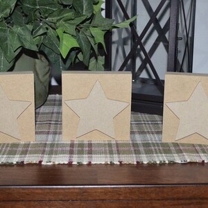 May include: Three wooden blocks with a star cutout on each side. The blocks are a light brown color and are arranged in a row on a plaid surface.