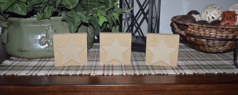 May include: Three wooden blocks with a star cutout on each side. The blocks are a light brown color and are arranged in a row on a plaid surface.