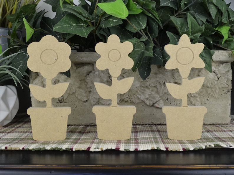 May include: Three unfinished wooden flower pot decorations. Each flower pot has a different flower design. The flower pots are all the same size and are made of a light brown wood.