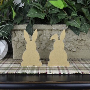 May include: Two unfinished wooden bunny shapes, one with a slightly raised ear, sitting on a plaid surface.