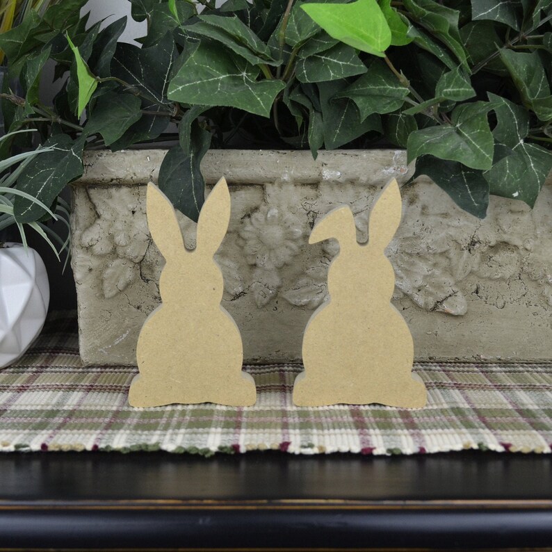 May include: Two unfinished wooden bunny shapes, one with a slightly raised ear, sitting on a plaid surface.