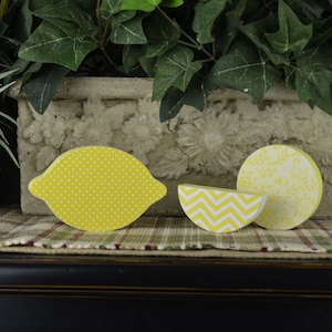 May include: Three wooden lemon-shaped ornaments with different patterns. The ornaments are yellow with white polka dots, white and yellow chevron, and white floral patterns.