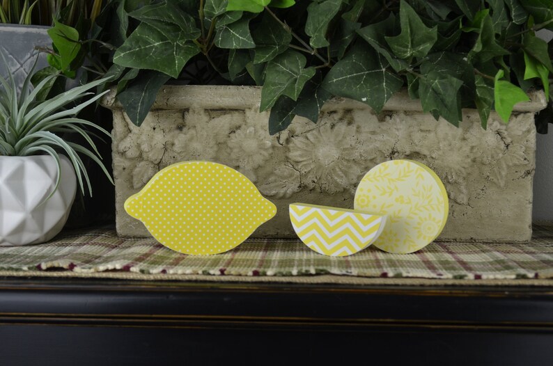 May include: Three wooden lemon-shaped ornaments with different patterns. The ornaments are yellow with white polka dots, white and yellow chevron, and white floral patterns.