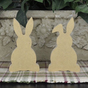 May include: Two unfinished wooden bunny shapes, one with its ears up and the other with one ear down, sitting on a plaid surface.