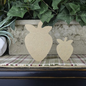 May include: Two wooden strawberry shapes, one larger than the other, are displayed on a plaid surface. The strawberries are a light brown color and have a simple, cutout design.