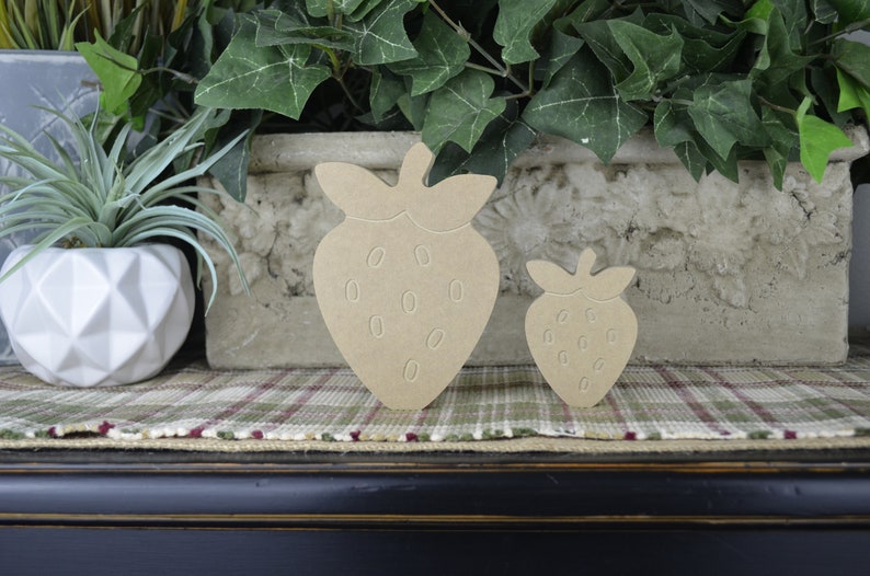 May include: Two wooden strawberry shapes, one larger than the other, are displayed on a plaid surface. The strawberries are a light brown color and have a simple, cutout design.