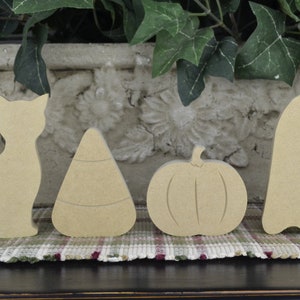 May include: Five unfinished wooden Halloween decorations: a cat, a candy corn, a pumpkin, a ghost, and a bat. The decorations are all in a natural wood color and are ready to be painted or decorated.