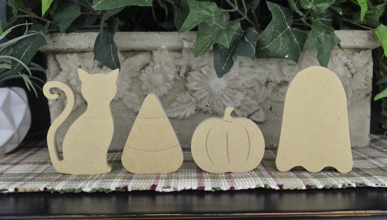 May include: Five unfinished wooden Halloween decorations: a cat, a candy corn, a pumpkin, a ghost, and a bat. The decorations are all in a natural wood color and are ready to be painted or decorated.
