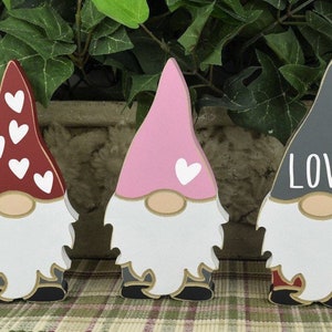 Op de afbeelding: Drie houten dwergen met geschilderde gezichten en hoeden. De dwerg links heeft een rode hoed met witte hartjes, de dwerg in het midden heeft een roze hoed met een wit hartje en de dwerg rechts heeft een grijze hoed met het woord "LOVE" in wit.