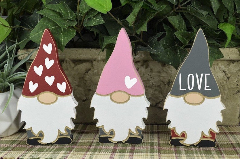Op de afbeelding: Drie houten dwergen met geschilderde gezichten en hoeden. De dwerg links heeft een rode hoed met witte hartjes, de dwerg in het midden heeft een roze hoed met een wit hartje en de dwerg rechts heeft een grijze hoed met het woord "LOVE" in wit.