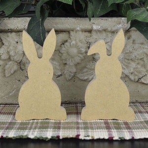May include: Two unfinished wooden bunny shapes, one facing forward and one facing backward, on a plaid surface.