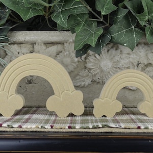 May include: Two wooden rainbow decorations with cloud accents. The rainbows are light brown and have a carved design. One is larger than the other. They are displayed on a plaid surface with greenery in the background.