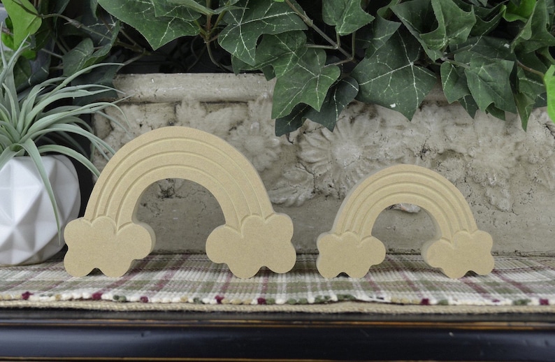 May include: Two wooden rainbow decorations with cloud accents. The rainbows are light brown and have a carved design. One is larger than the other. They are displayed on a plaid surface with greenery in the background.