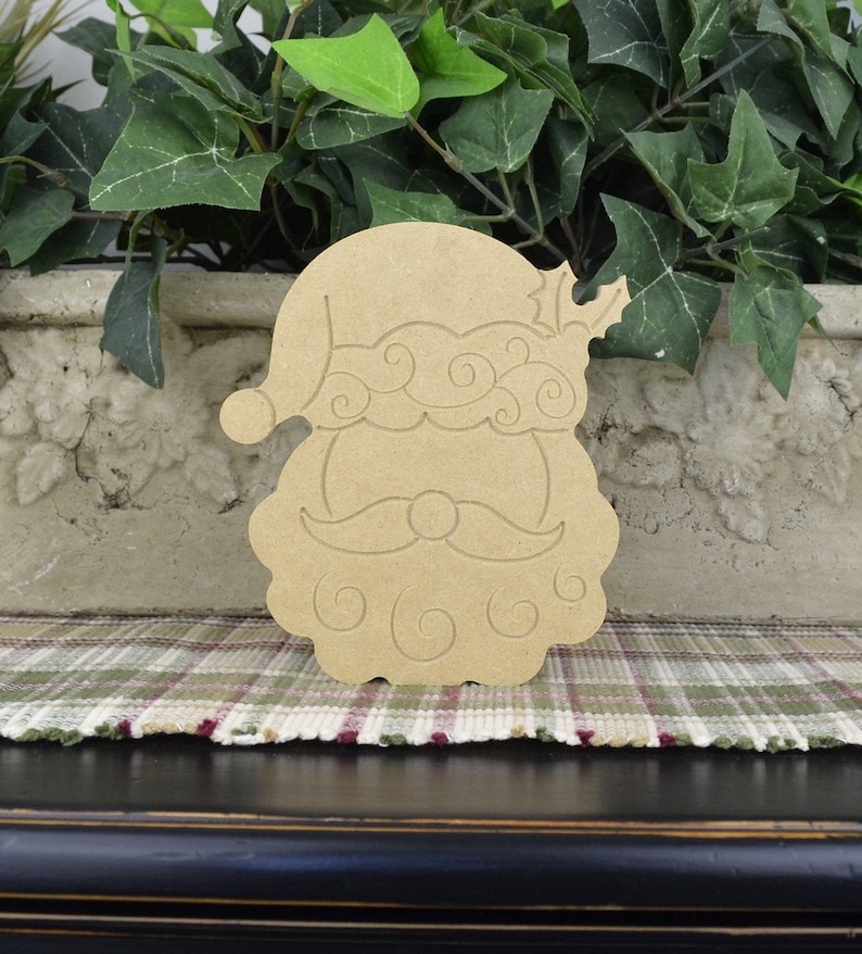 May include: Unfinished wooden cutout of Santa Claus's face with a curly beard and a hat. The cutout is a light brown color and has a simple design.