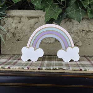 May include: A wooden rainbow decoration with pastel colors, including pink, blue, and purple arches, and white cloud accents. The rainbow is approximately 6 inches wide and 4 inches tall, with a gold outline.