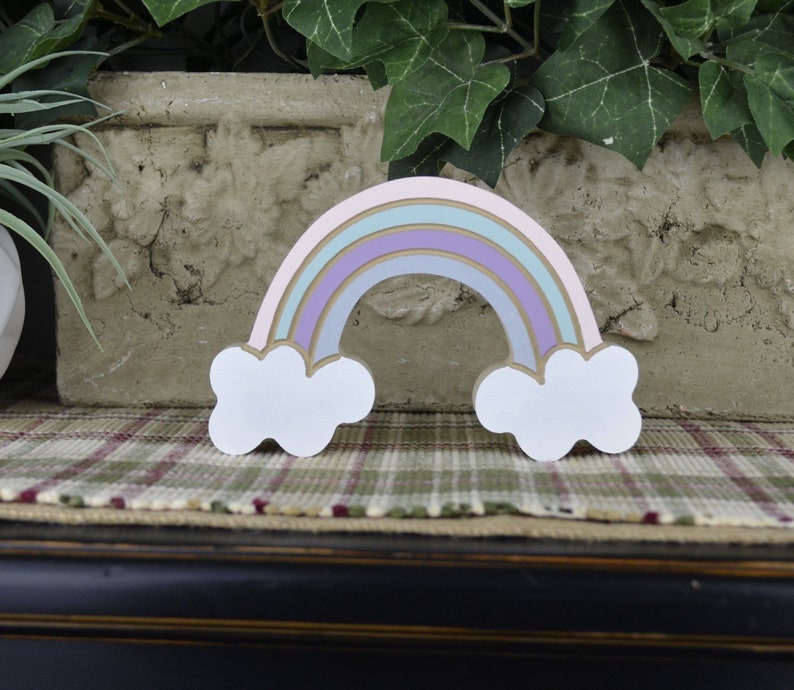 May include: A wooden rainbow decoration with pastel colors, including pink, blue, and purple arches, and white cloud accents. The rainbow is approximately 6 inches wide and 4 inches tall, with a gold outline.