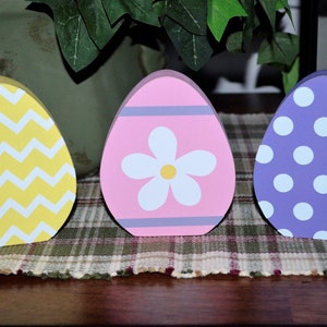 May include: Three wooden Easter egg decorations. One is yellow with white zigzags, the second is pink with a white flower, and the third is purple with white polka dots. They are displayed on a plaid surface.