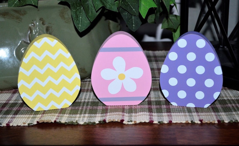 May include: Three wooden Easter egg decorations. One is yellow with white zigzags, the second is pink with a white flower, and the third is purple with white polka dots. They are displayed on a plaid surface.