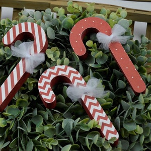 K&ouml;nnte beinhalten: Drei rote und wei&szlig;e Zuckerstangen-Ornamente mit wei&szlig;en T&uuml;llschleifen. Die Zuckerstangen sind mit Streifen, Punkten und einem Zickzackmuster verziert.