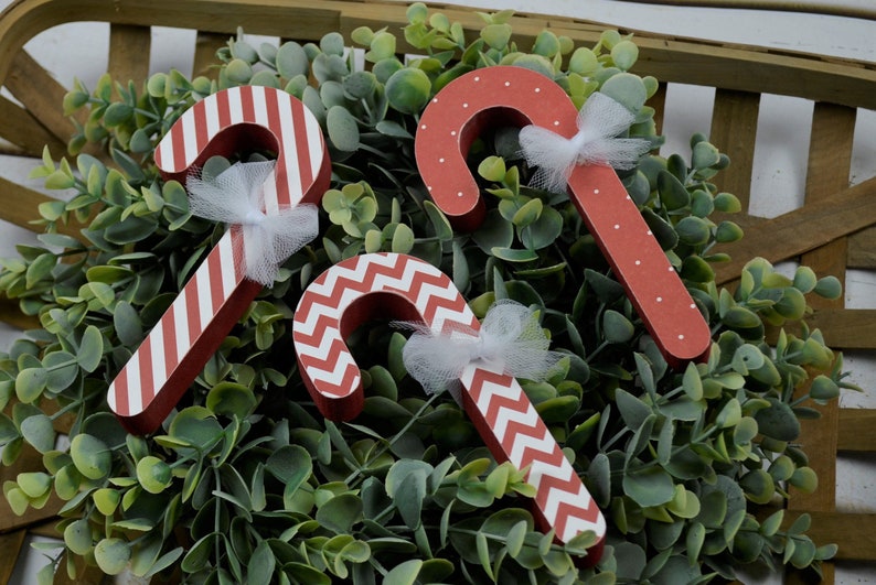 K&ouml;nnte beinhalten: Drei rote und wei&szlig;e Zuckerstangen-Ornamente mit wei&szlig;en T&uuml;llschleifen. Die Zuckerstangen sind mit Streifen, Punkten und einem Zickzackmuster verziert.