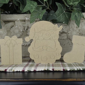 K&ouml;nnte beinhalten: Drei unfertige Weihnachtsornamente aus Holz: ein Weihnachtsmannkopf, ein Strumpf und eine Geschenkbox. Die Ornamente sind bereit zum Bemalen oder Dekorieren.