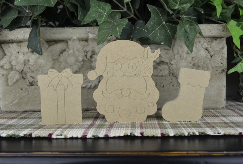 K&ouml;nnte beinhalten: Drei unfertige Weihnachtsornamente aus Holz: ein Weihnachtsmannkopf, ein Strumpf und eine Geschenkbox. Die Ornamente sind bereit zum Bemalen oder Dekorieren.