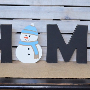 May include: Black wooden letters spelling "HOME" with a white snowman wearing a blue hat and scarf in the center.