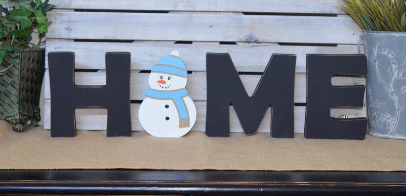 May include: Black wooden letters spelling "HOME" with a white snowman wearing a blue hat and scarf in the center.