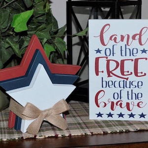 May include: A wooden sign with the text "land of the free because of the brave" in red and blue lettering. There are five stars below the text. The sign is sitting on a plaid table runner. A three-dimensional star made of wood painted red, white, and blue is in the foreground.