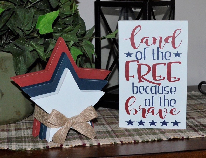 May include: A wooden sign with the text "land of the free because of the brave" in red and blue lettering. There are five stars below the text. The sign is sitting on a plaid table runner. A three-dimensional star made of wood painted red, white, and blue is in the foreground.
