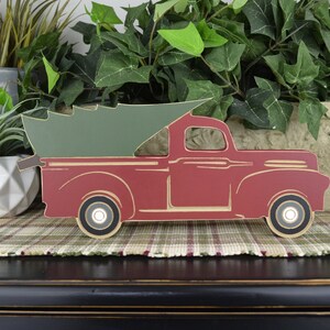 May include: A red wooden truck with a green Christmas tree in the bed. The truck is on a plaid surface.