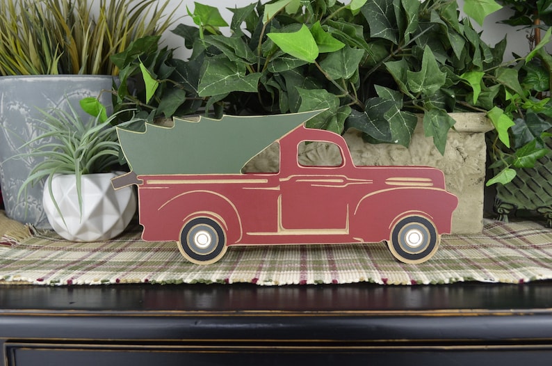 May include: A red wooden truck with a green Christmas tree in the bed. The truck is on a plaid surface.