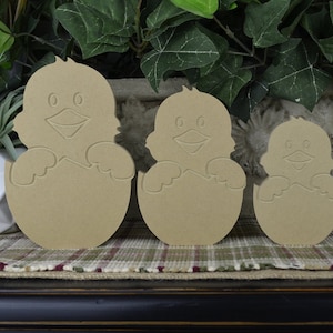 May include: Three wooden cutouts of chicks hatching from eggs. The chicks are in different sizes, with the largest chick being about 6 inches tall. The cutouts are made of unfinished wood and are ready to be painted or stained.