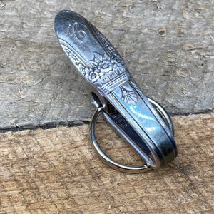 May include: A silver spoon keychain with a floral design and a ring attached. The spoon has an inscription on the top.