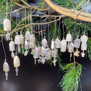 May include: A collection of silver Christmas ornaments in the shape of bells, each with a different design. The ornaments are hanging from a branch of a pine tree.