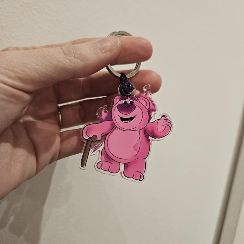 Puede incluir: Llavero de oso de dibujos animados rosa con un bast&oacute;n, mostrando una expresi&oacute;n amigable. El llavero est&aacute; hecho de acr&iacute;lico transparente y tiene un anillo de metal plateado.