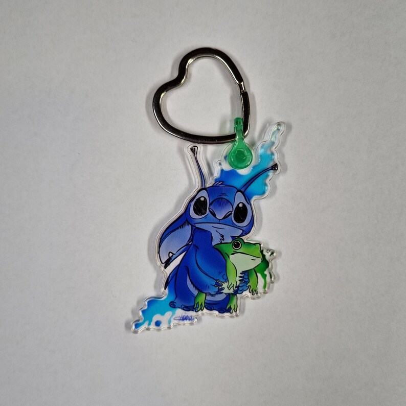 Puede incluir: Llavero con forma de coraz&oacute;n plateado con un personaje azul Stitch sosteniendo una rana verde.