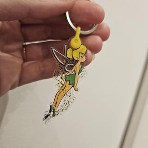 Puede incluir: Un llavero de acr&iacute;lico transparente con Campanilla, un hada de Disney, con alas verdes y un vestido verde. Ella est&aacute; volando con brillo amarillo a su alrededor.
