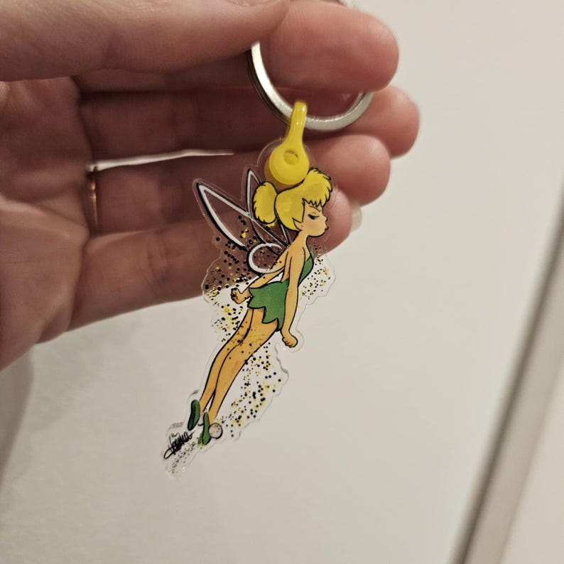 Puede incluir: Un llavero de acr&iacute;lico transparente con Campanilla, un hada de Disney, con alas verdes y un vestido verde. Ella est&aacute; volando con brillo amarillo a su alrededor.