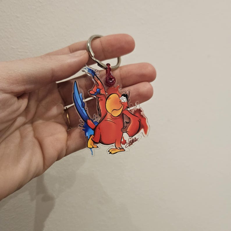 Puede incluir: Un llavero de loro de dibujos animados rojo y azul con fondo blanco. El loro sostiene un micr&oacute;fono y tiene una expresi&oacute;n de sorpresa en su rostro.