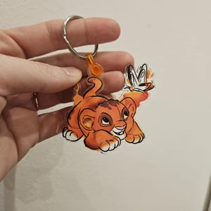 Puede incluir: Un llavero de acr&iacute;lico transparente con un cachorro de le&oacute;n de dibujos animados con pelaje naranja y una mariposa. El llavero est&aacute; unido a un anillo de metal plateado.